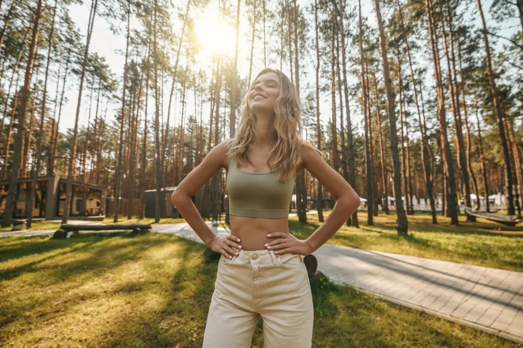 Gebrauchsanleitung - Inner balance. Smiling pretty young woman with closed eyes standing with hands on her belt in nature under rays of autumn sun
